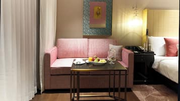 A hotel room with a pink sofa, a small table with fruits and a glass, a bed with white bedding, and a decorative wall piece.