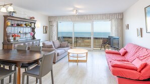 A living room with a sofa, a dining table with chairs, a coffee table, a TV, and a view of the sea through a sliding glass door. - Bright Apartment on the 7th Floor (Middelkerke)