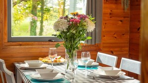 A dining table set with plates, bowls, and glasses, a vase with flowers, and a window with a view of greenery. - Lodge 6 - Hot Tub Lodge by Interhome (Launceston)