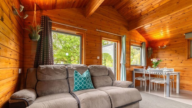A cozy cabin interior with a sofa, dining table, and large windows. - Lodge 6 - Hot Tub Lodge by Interhome (Launceston)