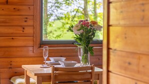 A wooden cabin interior with a dining table set for two, a vase with flowers, and a window offering a view of trees. - Lodge 4 Hot Tub by Interhome (Launceston)