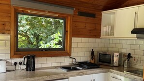 A kitchen with wooden cabinets, a window with a view of greenery, a coffee maker, a microwave, and a dishwasher. - Lodge 4 Hot Tub by Interhome (Launceston)