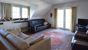 A spacious living room with a black leather sofa, a beige sofa, a coffee table, and a television. There is a large window with curtains, a floor lamp, and a decorative wall piece. - Ter Streep 0201 Apartment Near the Beach (De Haan)