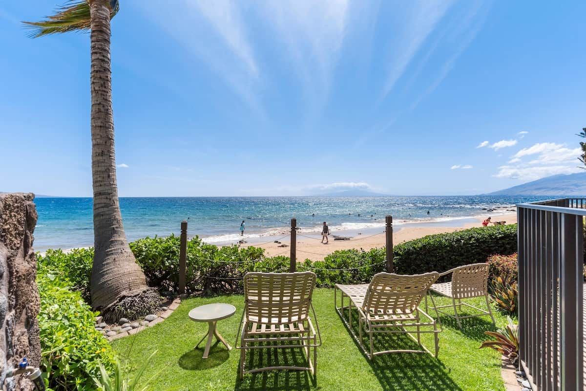 A terrace with two chairs and a table overlooking a beach with palm trees and clear skies.