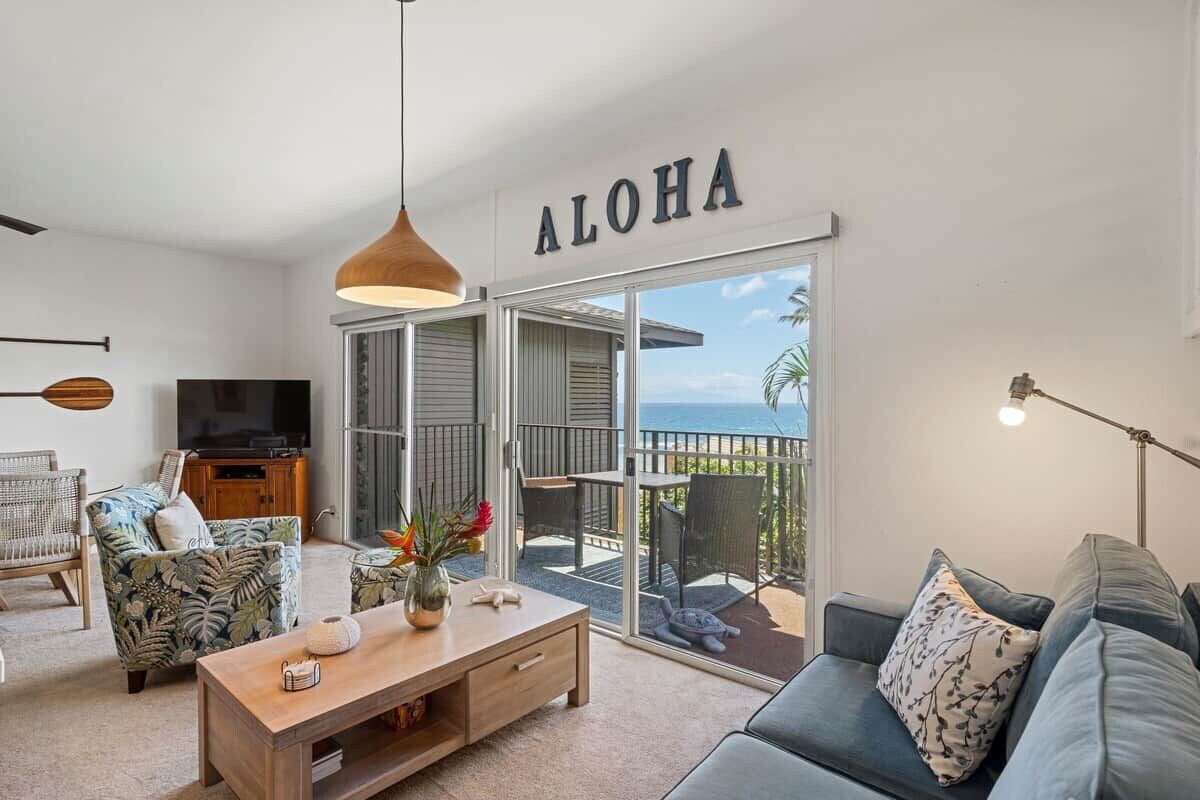 Sala de estar com sofá, mesa de centro e televisão. Um letreiro com a palavra 'ALOHA' está acima de uma porta de vidro deslizante que dá para uma varanda.
