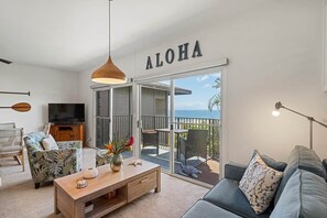 Una sala de estar con sofá, mesa de centro y televisor. Un cartel con la palabra 'ALOHA' está sobre una puerta corrediza de vidrio que da a un balcón.