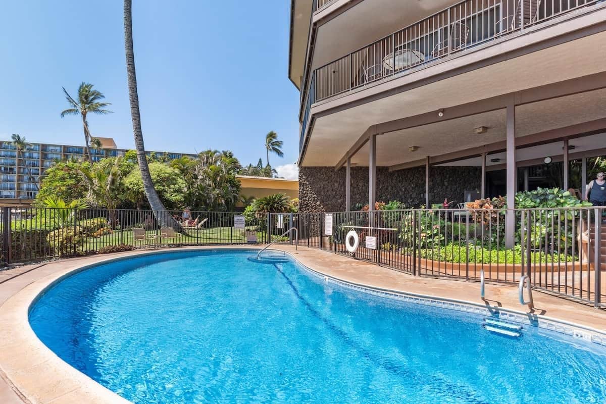 A swimming pool with a curved edge, surrounded by a fence, and a building with a balcony in the background.