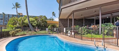 Una piscina de borde curvo, rodeada por una valla, y un edificio con balcón al fondo.