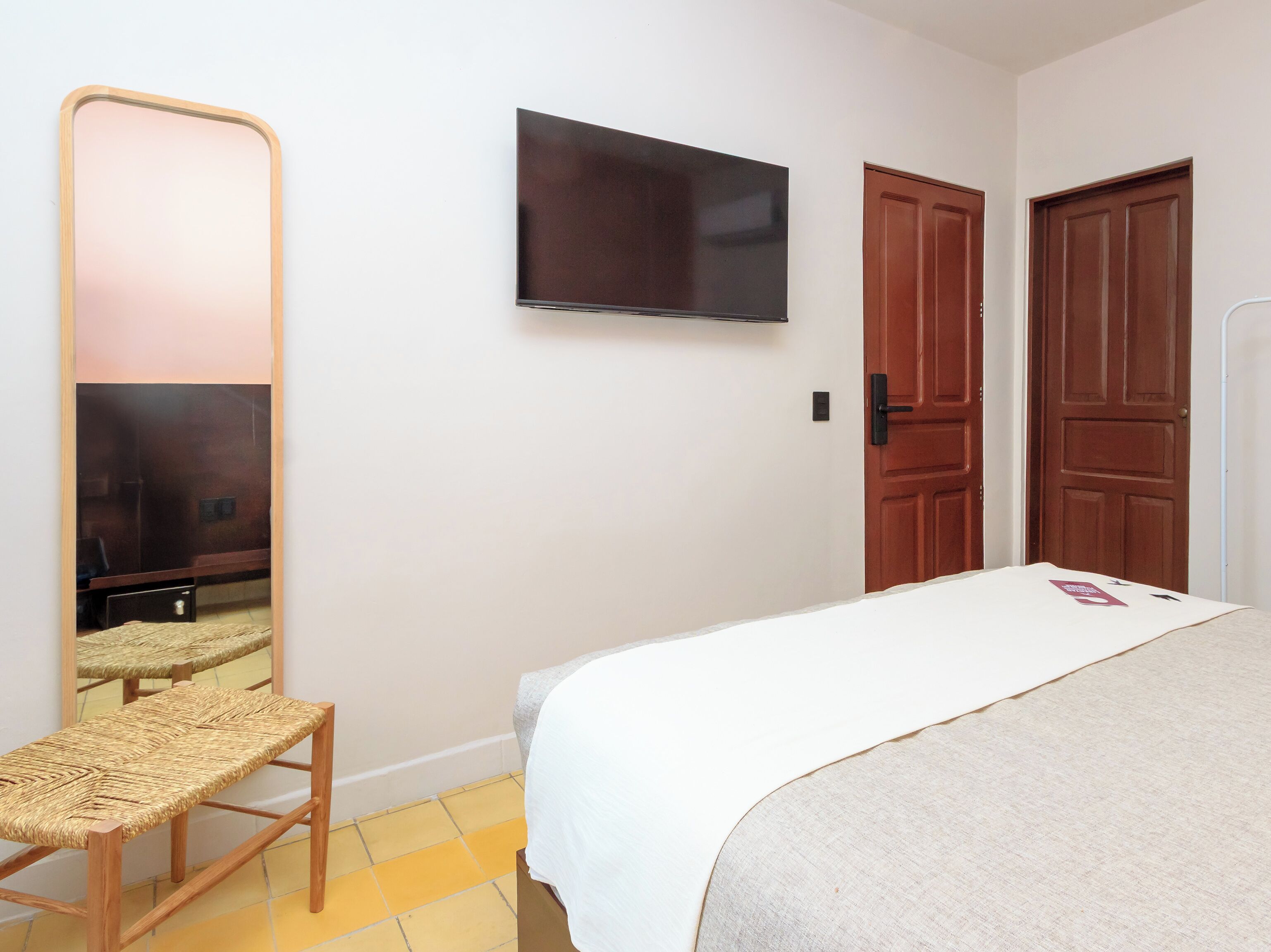 Un dormitorio con una cama, una silla de madera, un espejo grande, un televisor y dos puertas de madera.