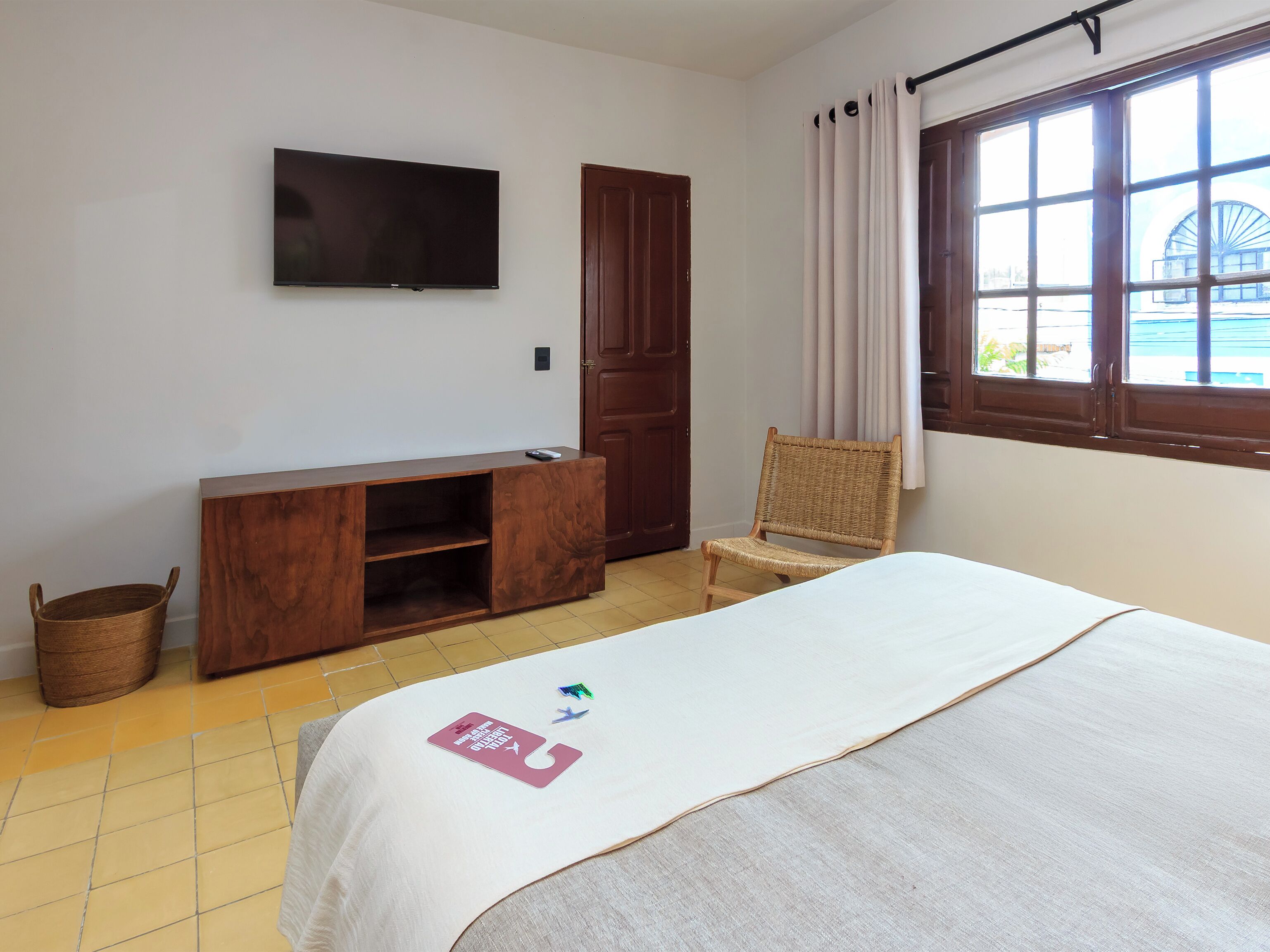 Quarto com uma cama, televisão na parede, armário de madeira, cadeira de vime e janela com cortinas.
