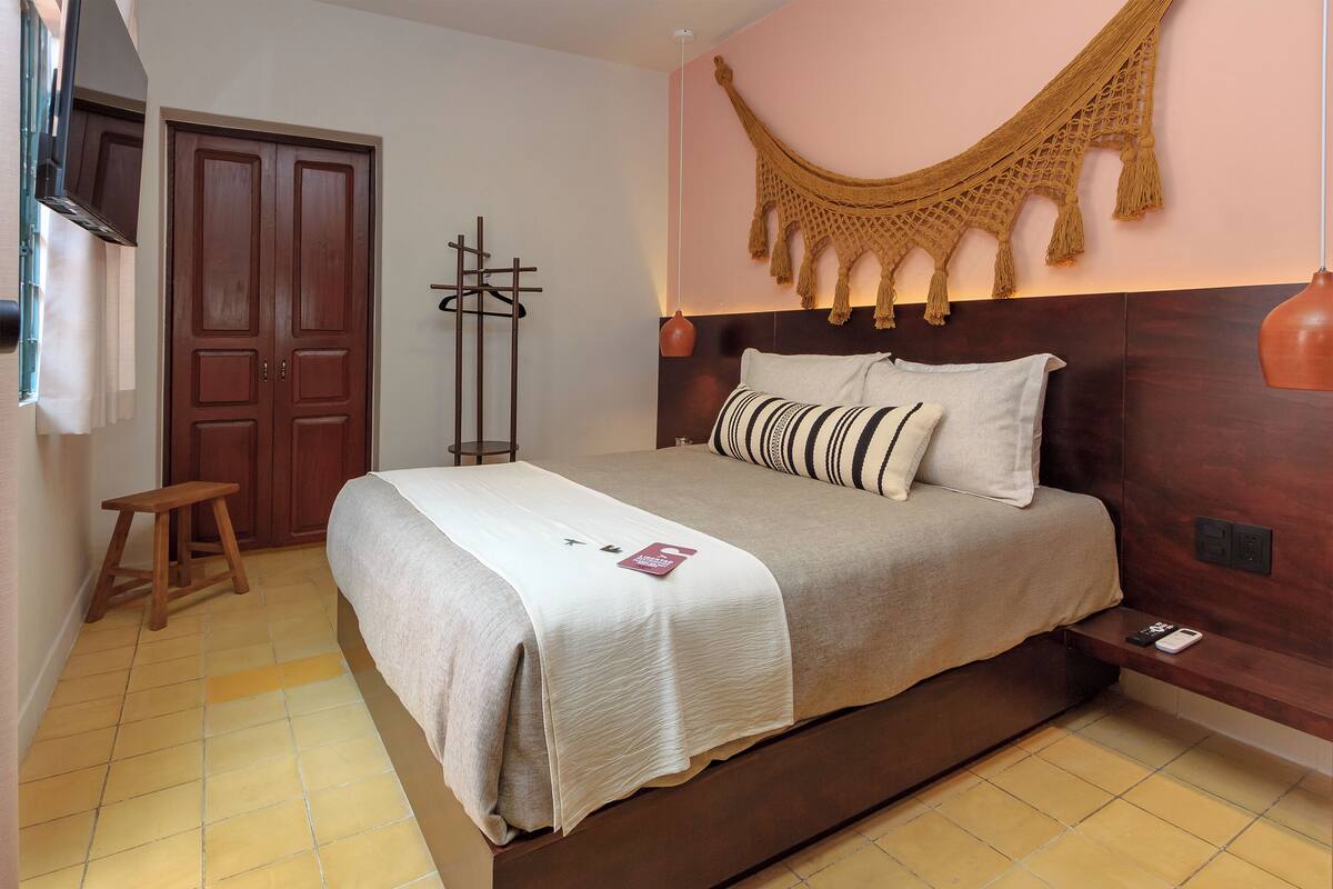 Een slaapkamer met een groot bed, houten hoofdeinde, een macramé wanddecoratie en een klein houten krukje.