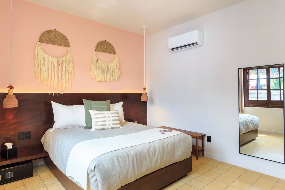 Een netjes opgemaakt slaapkamer met een bed, nachtkastjes, een wandairconditioner en een grote spiegel die de kamer weerspiegelt.