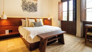 A neatly arranged bedroom with a wooden headboard, a hanging wall decoration, a bedside table, and a large window with wooden shutters. - Hotel Libertad 1416 (Guadalajara)