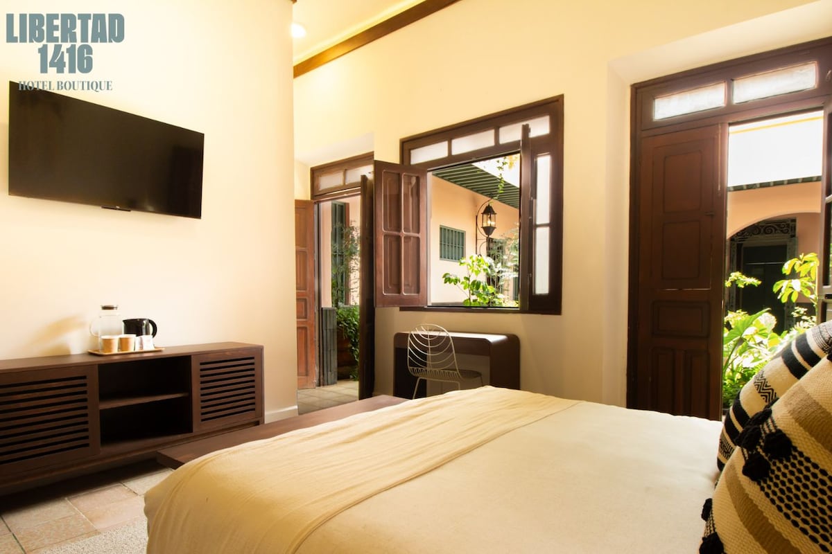Habitación de hotel con cama, televisión montada en la pared, ventana con vistas a zonas verdes y puerta que da a un balcón.