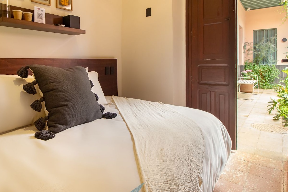 Una habitación de hotel con una cama, cabecera de madera, estante con marcos de fotos y una puerta que da a un área exterior.