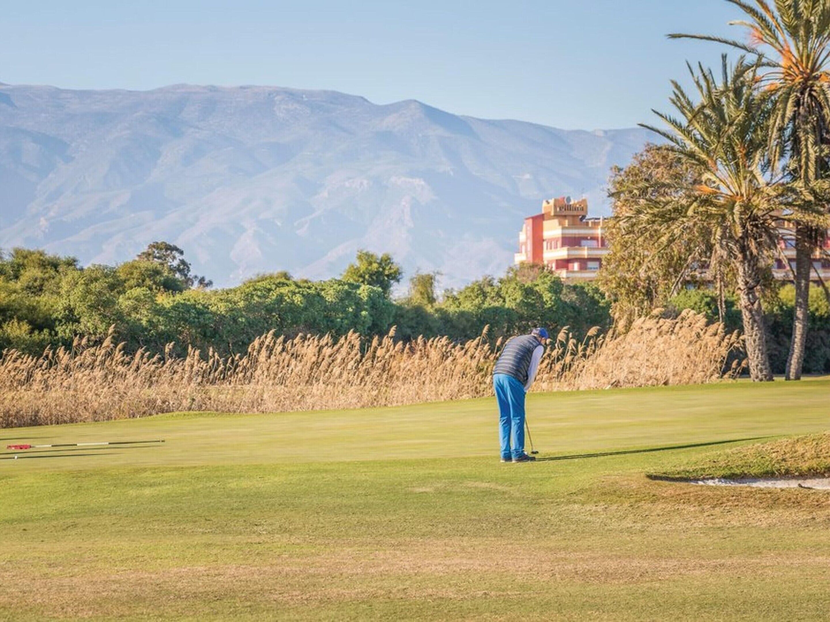 Un parcours de golf avec une chaîne de montagnes en arrière-plan, un bâtiment à la façade rouge et jaune, et des palmiers.