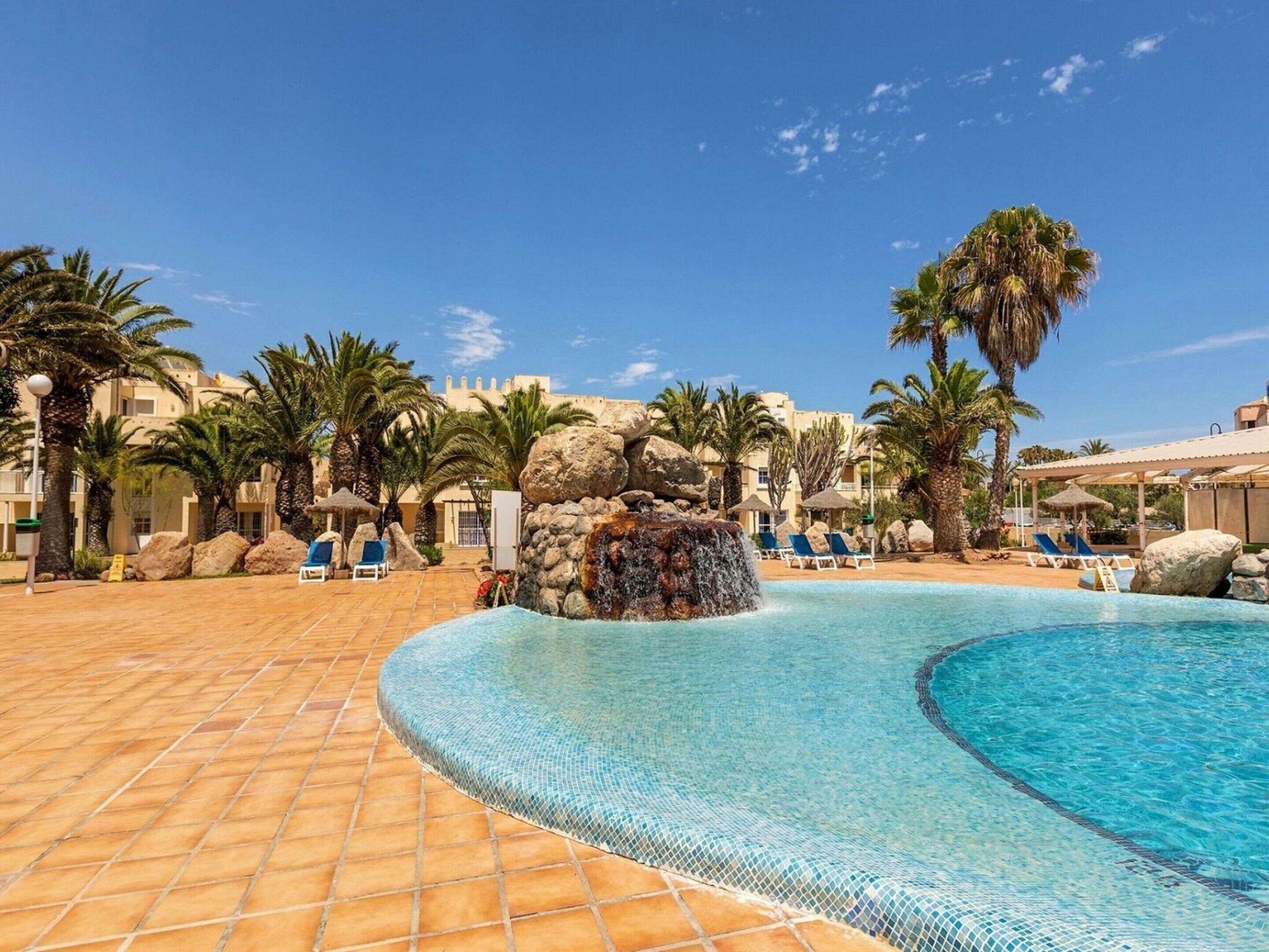Ein Poolbereich eines Resorts mit klarem, blauem Pool, Liegestühlen, Palmen und einem Wasserfall-Element mit Felsen.