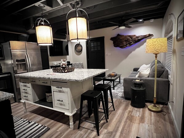 A modern kitchen with a central island, black bar stools, a dining table, and a sofa in a living area.