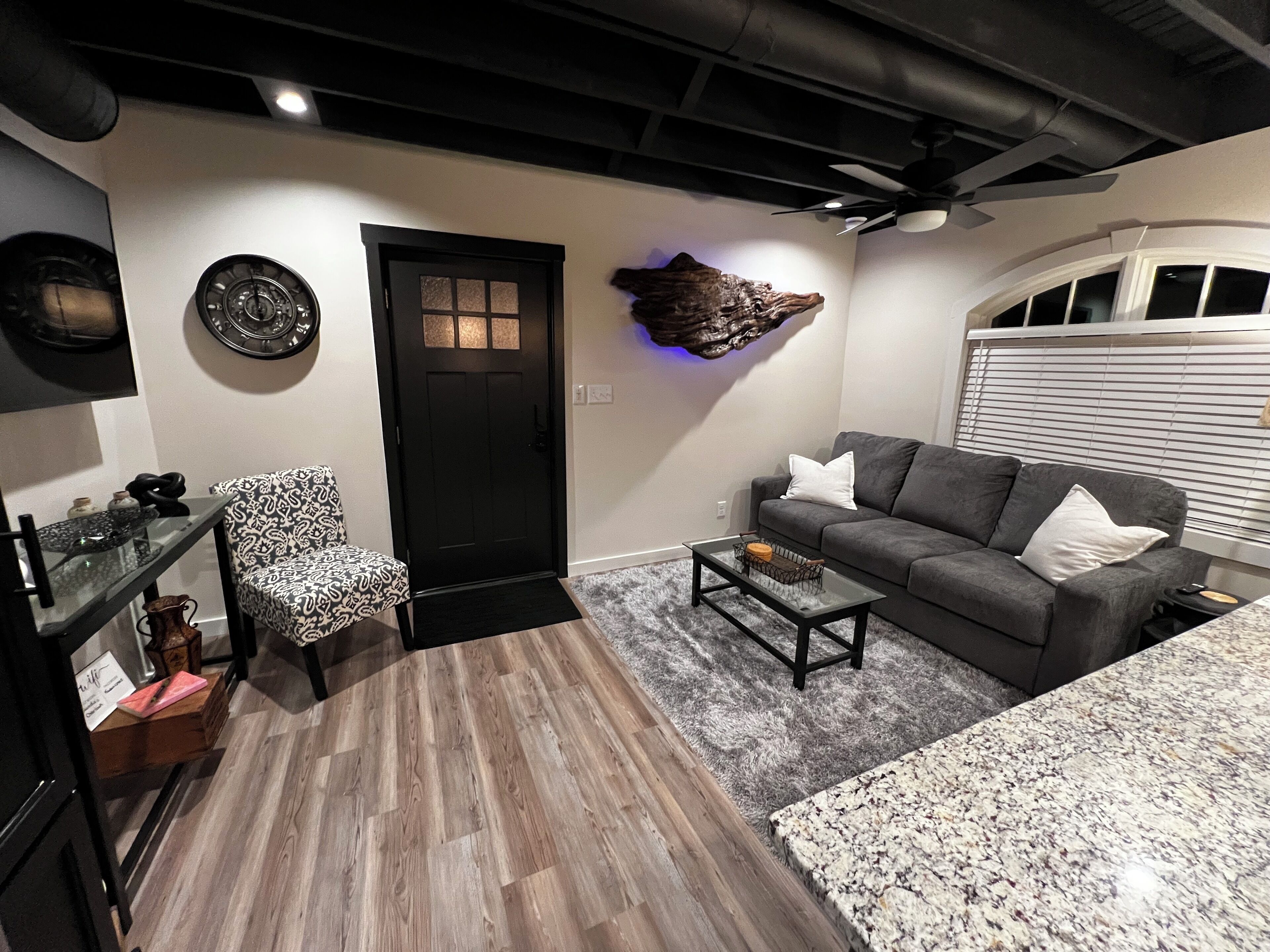 Un salon moderne avec une porte sombre, un canapé gris, un fauteuil à motifs, une table basse et un ventilateur de plafond.