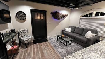Un salon moderne avec une porte sombre, un canapé gris, un fauteuil à motifs, une table basse et un ventilateur de plafond.