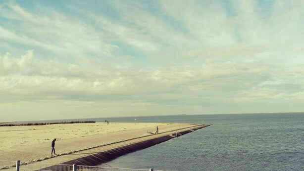 Une plage avec une longue jetée qui s’avance dans la mer, une rive sablonneuse et quelques personnes qui se promènent le long de la plage.