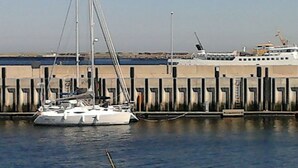 Un port de plaisance où un voilier est amarré près d’un quai, avec un grand navire en arrière-plan et une bouée jaune flottant sur l’eau.