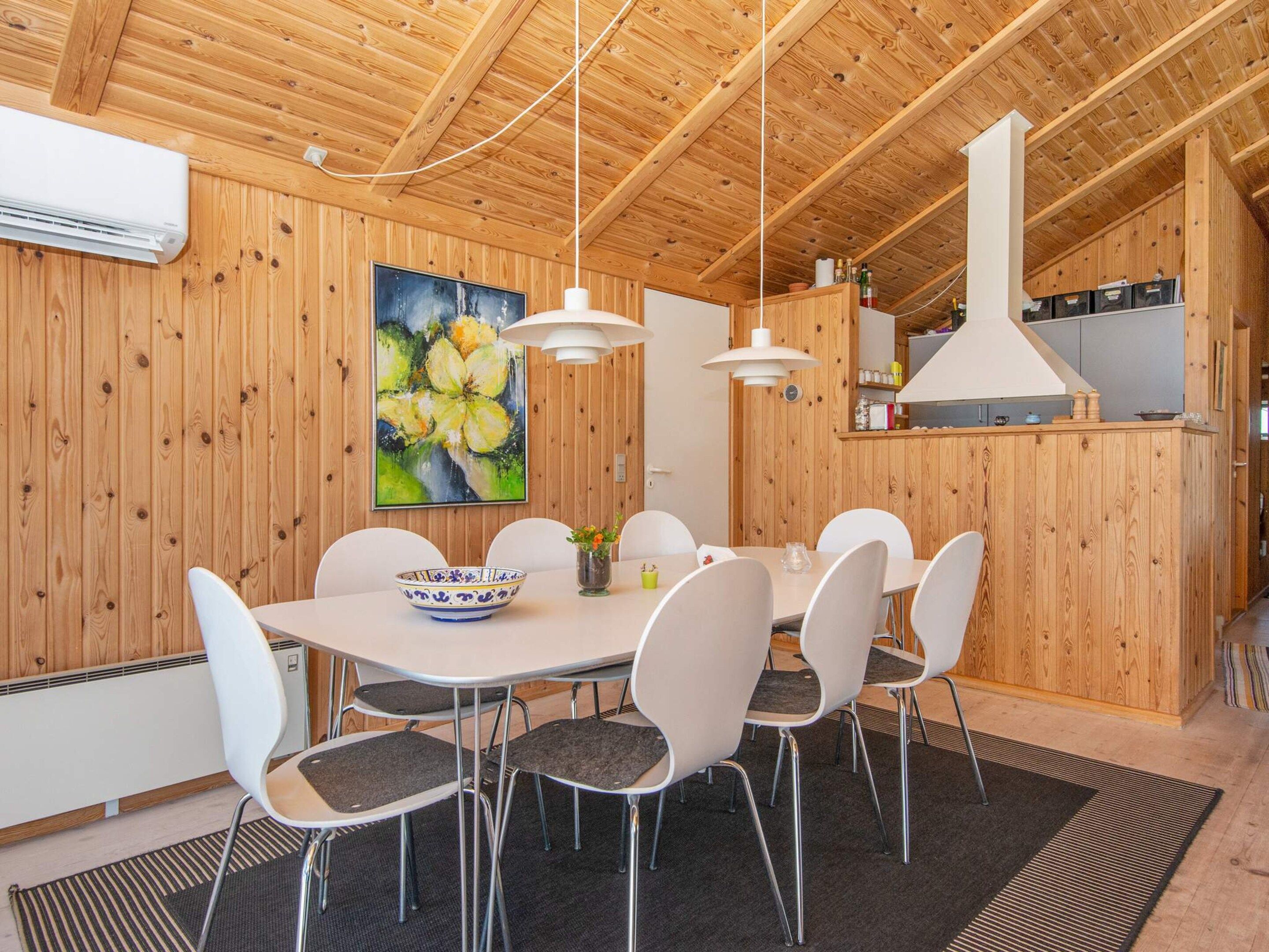 A wooden-paneled room with a dining table, chairs, a painting, and a kitchen area.