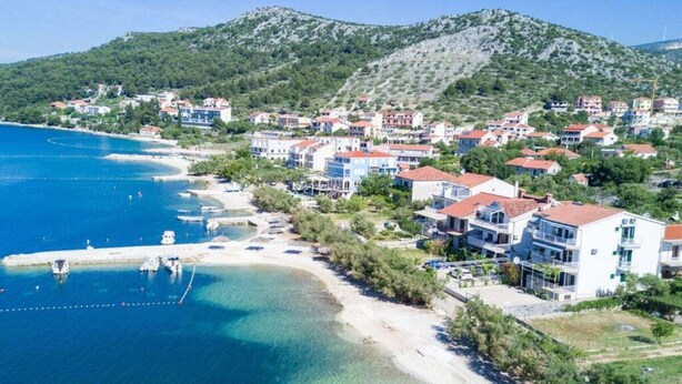 Une ville côtière avec des bâtiments qui bordent la plage, une eau d’un bleu limpide et un paysage vallonné en arrière-plan.