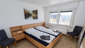 A modern bedroom with a wooden bed, two bedside tables, a chair, and a large window with a view. - Fewo Freiken 28 Comfortable Holiday Residence (Arnsberg)