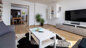 A modern living room with a white sofa, a white coffee table, a flat-screen TV, and a dining area with a wooden table and chairs. - Fewo Freiken 28 Comfortable Holiday Residence (Arnsberg)