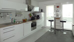 Cocina moderna con armarios blancos, electrodomésticos de acero inoxidable, microondas y cafetera. Hay dos taburetes de bar y una ventana con persianas.