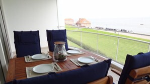 Un balcón con una mesa de madera dispuesta para dos, sillas con cojines azules y una vista despejada del mar y edificios al fondo.