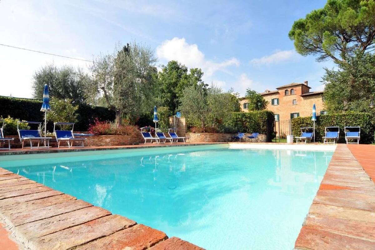 A clear swimming pool with lounge chairs and umbrellas, surrounded by trees and a building.