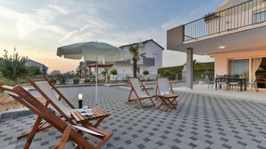 Et udendørs opholdsområde med træloungestole og borde, en overdækket terrasse og en bygning med balkon.