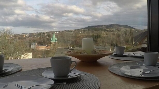 A breakfast table with plates, cups, and a candle, overlooking a cityscape with buildings and a hill. - Uitzicht op Droomlocatie (Annaberg-Buchholz)