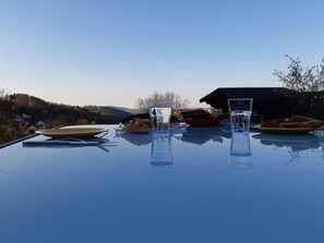 Una mesa con vasos de agua, un plato de comida y un tazón, colocada al aire libre con vista a una casa y colinas.