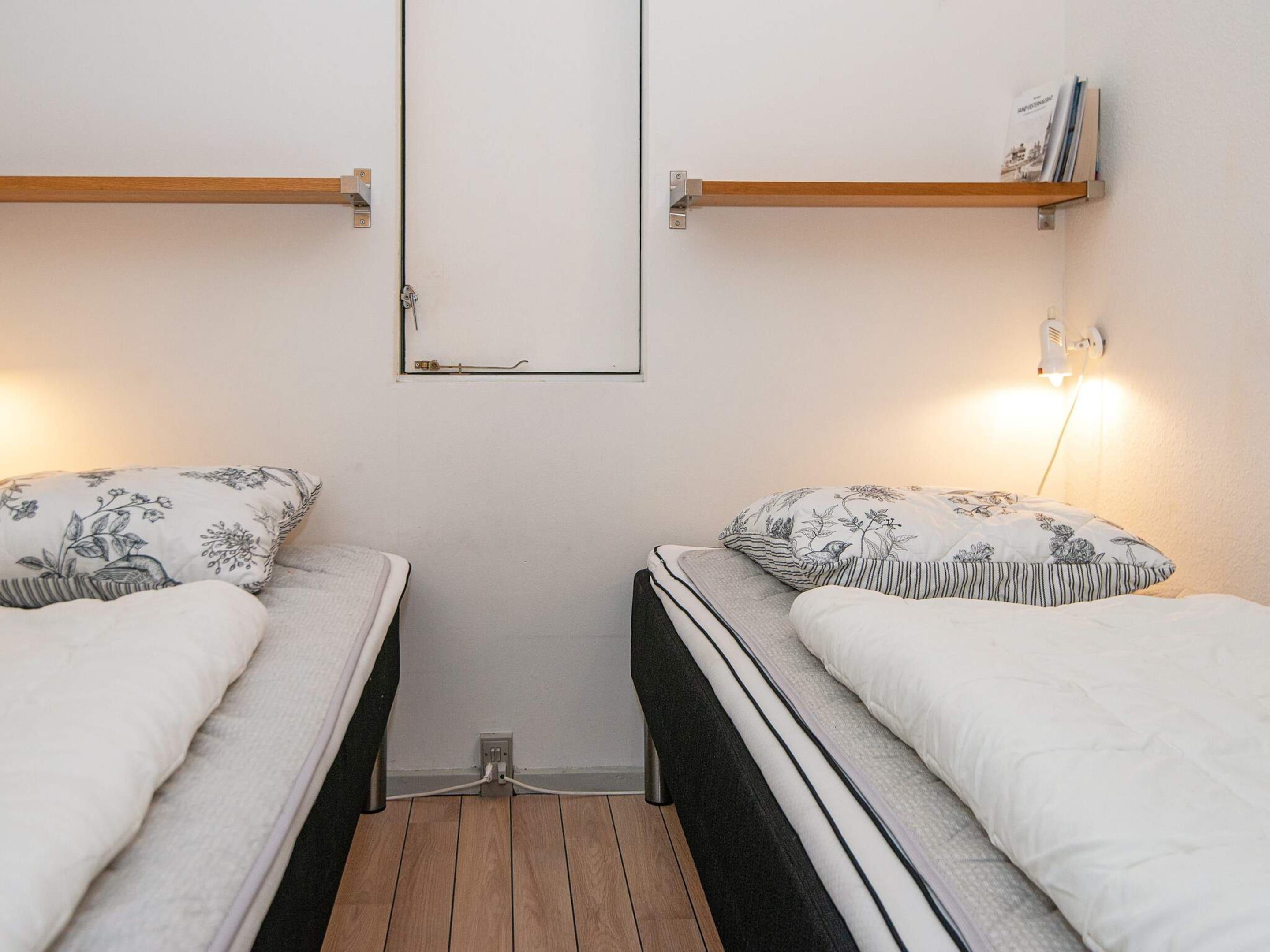 A small, compact room with two single beds, each with a wooden headboard and a floral-patterned pillow. There is a wall-mounted shelf on the left and a wall-mounted lamp on the right.
