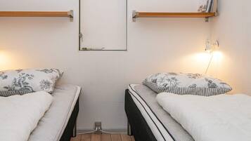 A small, compact room with two single beds, each with a wooden headboard and a floral-patterned pillow. There is a wall-mounted shelf on the left and a wall-mounted lamp on the right.