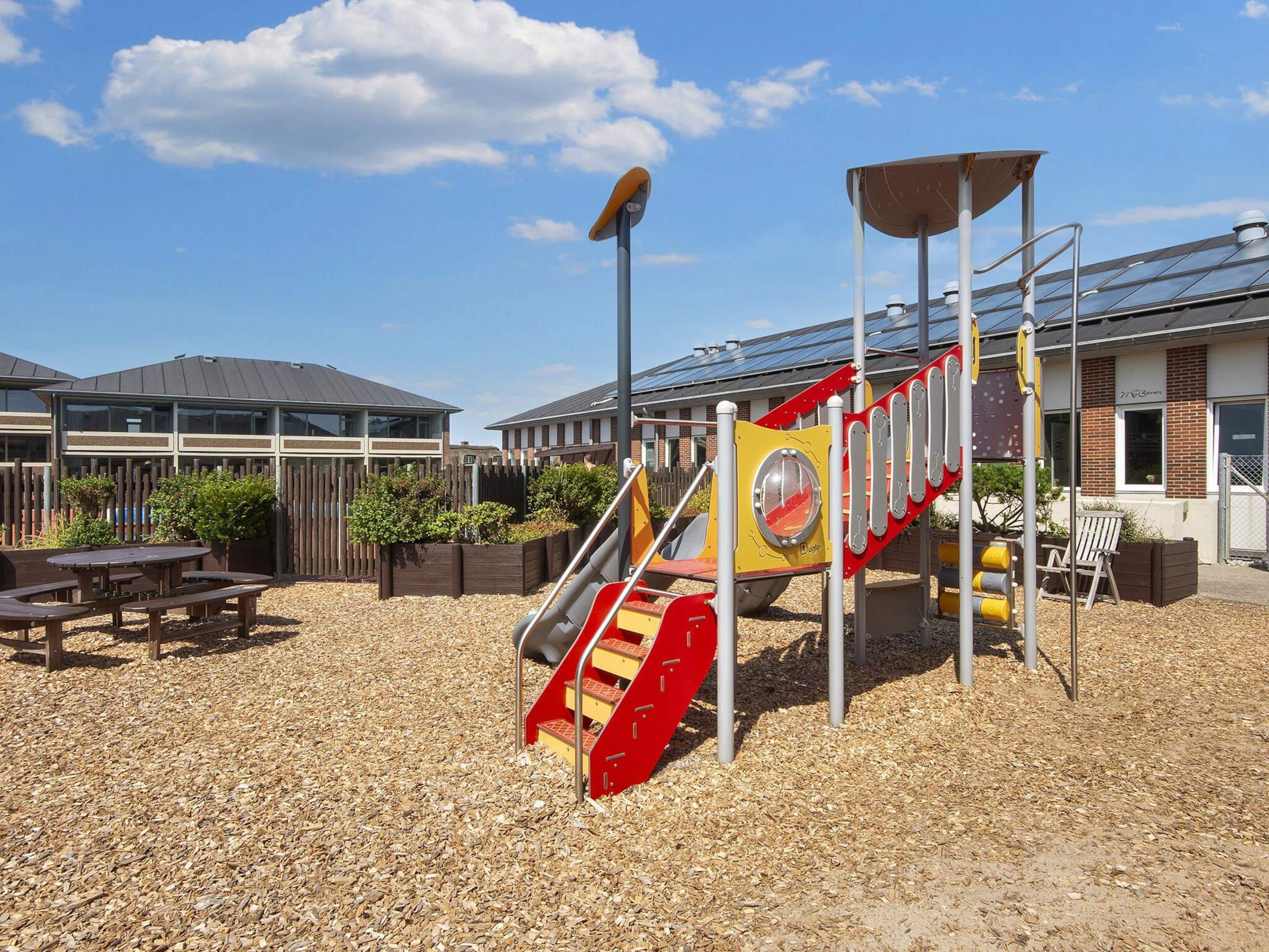 A playground with a slide and climbing structure, surrounded by a wooden fence and picnic tables.