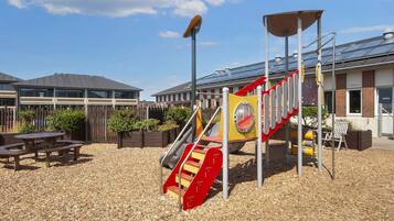 A playground with a slide and climbing structure, surrounded by a wooden fence and picnic tables.