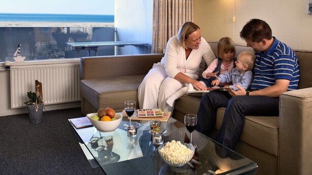 Eine Familie sitzt auf einer Couch mit Blick auf den Ozean, ein Glastisch mit Snacks und Getränken sowie ein Fenster mit Vorhängen.