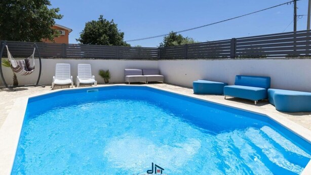 Une piscine entourée de chaises longues blanches et de canapés bleus, le tout bordé d’un mur blanc et d’une clôture noire.