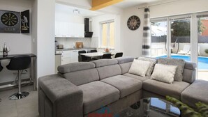 Un salon moderne avec un canapé d’angle gris, un coin repas avec une table et des chaises, une cuisine avec des meubles blancs et une grande fenêtre donnant sur une piscine.