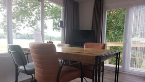A dining area with a wooden table and chairs, a television, and large windows with curtains. - Cosy Holiday Home in Appelscha With a Great View (Appelscha)