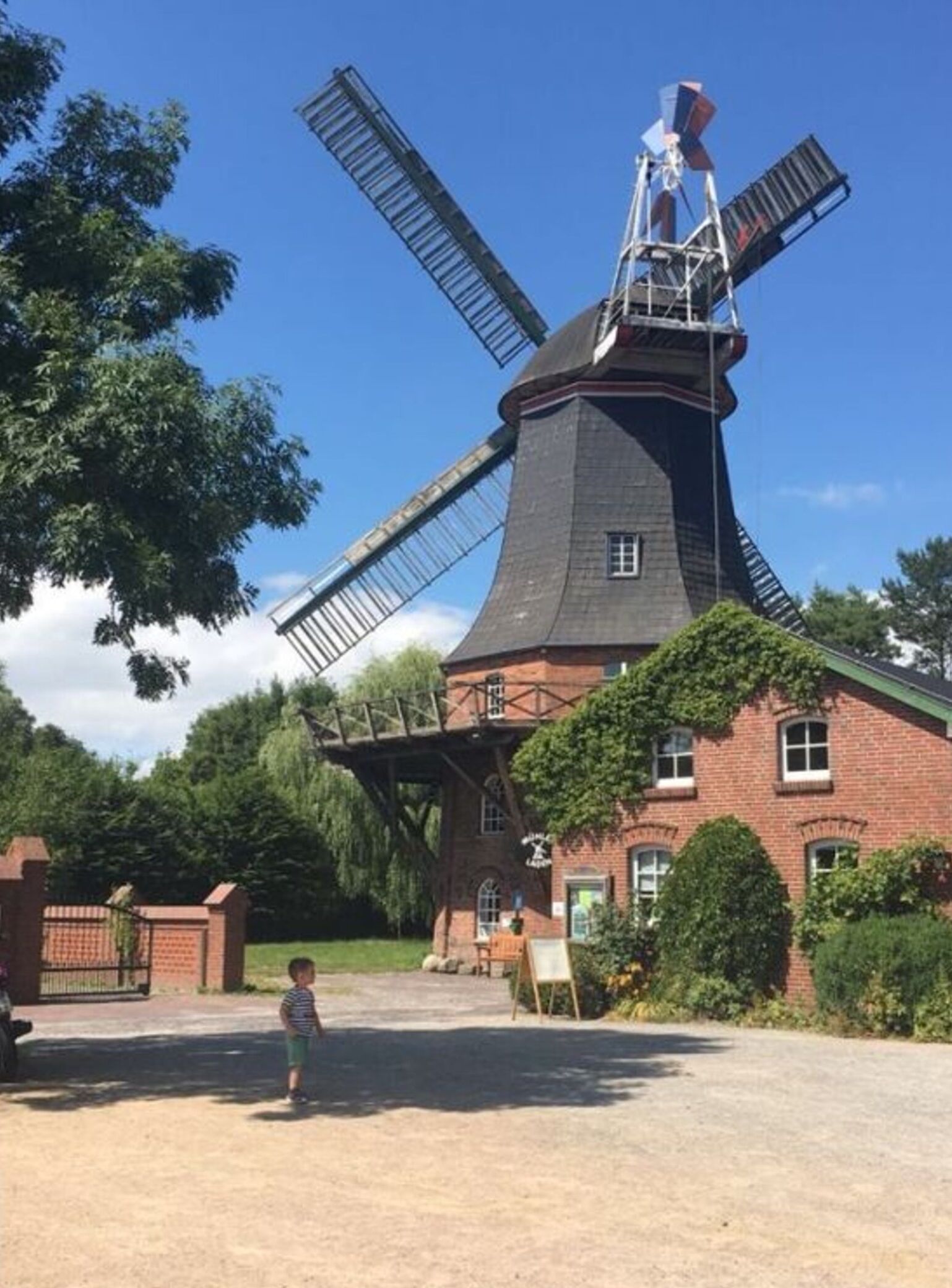 A windmill with a brick base and a metal structure, surrounded by greenery and a clear sky.