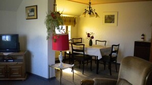 Una sala de estar con televisor, una mesa de comedor con sillas, un sofá y una mesita con un florero.