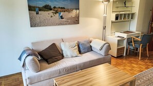 A living room with a beige sofa, a wooden coffee table, a desk with a chair, and a large painting of a beach scene. - Kersten by Interhome (Essen)