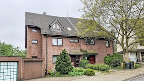 A two-story brick house with a sloped roof, a front garage, and a small front yard with a tree and shrubs. - Kersten by Interhome (Essen)