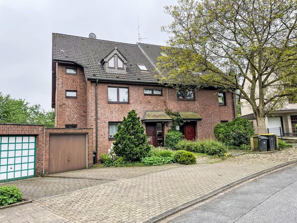 A two-story brick house with a sloped roof, a front garage, and a small front yard with a tree and shrubs. - Kersten by Interhome (Essen)