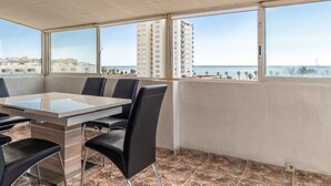 A modern dining area with a glass table and black chairs, large windows offering a view of the sea and buildings. - Apartment Jazmin by Interhome (Salou)